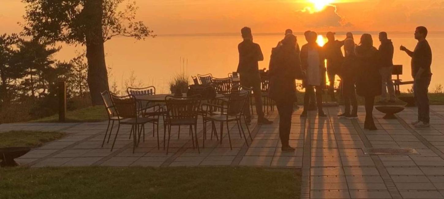Gruppe mennesker står på terrassen ved Bautahøj Hotel & Konferencecenter og nyder en smuk solnedgang over vandet.
