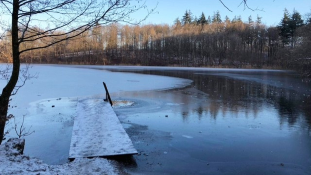 Vinterstemning ved Avnsø med delvist frossen sø, sneklædt bred, badebro og skov i baggrunden.