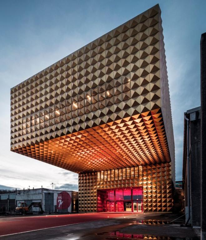 Gylden, kubeformet facade på Ragnarock med markant udhæng og rødlyst indgang i aftenskumring.