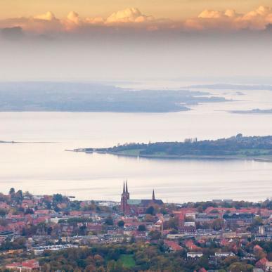 Udsigt over Roskilde med Roskilde Domkirke i forgrunden og Roskilde Fjord omgivet af øer og landskab i baggrunden under en himmel med skyer i aftensol.