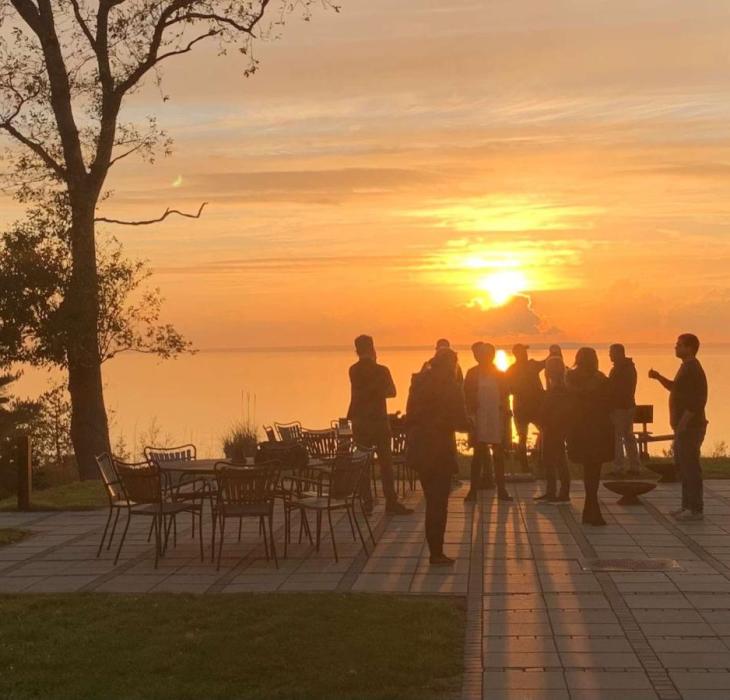 Gruppe mennesker står på terrassen ved Bautahøj Hotel & Konferencecenter og nyder en smuk solnedgang over vandet.
