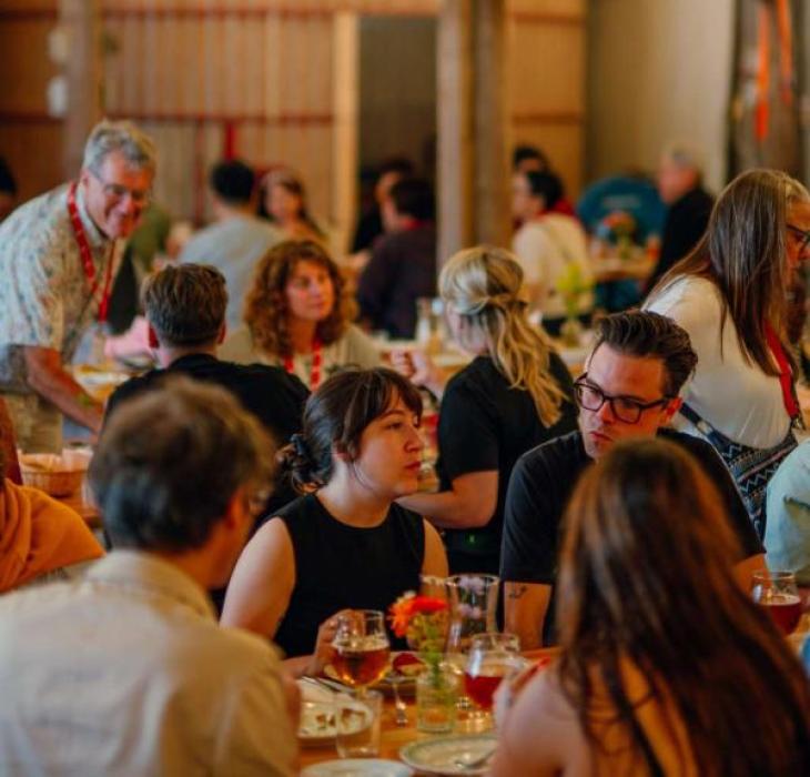 Gæster sidder ved langborde i en rustik lade hos Herslev Bryghus, omgivet af farverige papirdekorationer og hyggelig belysning.