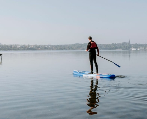 Mand der står på SUP-board med padel i hånden på Roskilde Fjord. Taget ved Vigen Strandpark. Roskildes skyline er i baggrunden, hvor man kan se