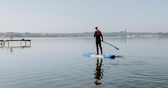 Mand der står på SUP-board med padel i hånden på Roskilde Fjord. Taget ved Vigen Strandpark. Roskildes skyline er i baggrunden, hvor man kan se 